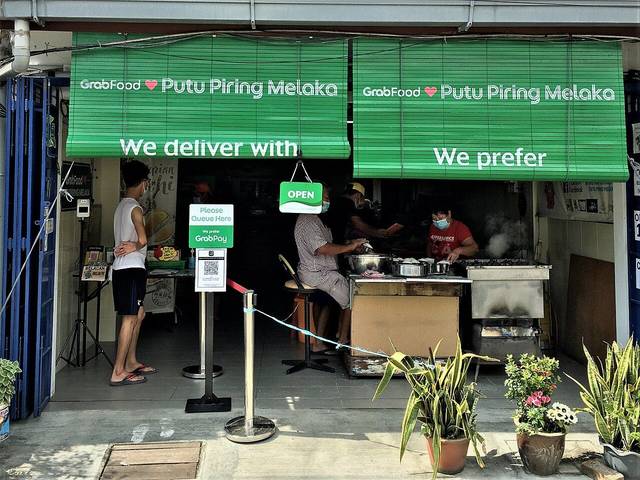 Shopfront of Putu Piring Melaka on Jalan Tengkera with green blinds, a queue barrier and staff steaming putu piring, photographed in 2021