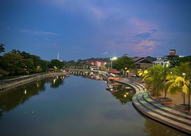 The Melaka River at dusk with a lit riverside walkway, palm trees and the old town reflected in the water